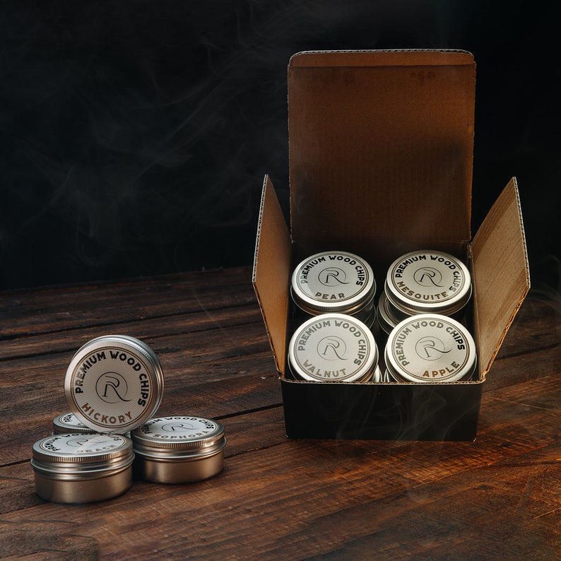 May include: A set of ten round metal tins containing wood chips for smoking food. The tins are labeled with the type of wood chip, including Hickory, Pear, Mesquite, Walnut, and Apple. The tins are arranged in a box with a black interior.