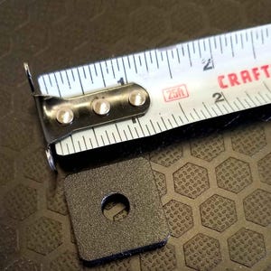 May include: A black square metal bracket with a central hole, next to a Craftsman 25-foot (7.62 m) measuring tape. The tape displays inch and foot markings.