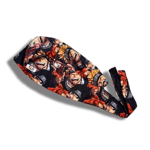 Op de afbeelding: Zwarte durag met een herhaald patroon van anime-gezichten in oranje, geel en zwart. De durag heeft een lange staart en een brede band. De tekst "NRS BAL" is zichtbaar op de durag.