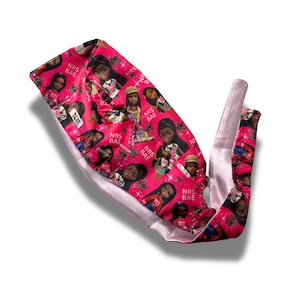 May include: A pink satin head wrap featuring a repeating pattern of dolls and the text "NRS BAE". The head wrap has a light pink satin lining and ties.