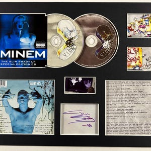 Puede incluir: CD de edición especial "The Slim Shady LP" de Eminem enmarcado. La presentación incluye el CD, la carátula del álbum y un autógrafo firmado. La carátula del álbum presenta una imagen en tonos azules de Eminem. También se incluye arte y texto adicional.