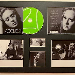 Puede incluir: Una foto enmarcada de la portada del álbum 21 de Adele. La portada del álbum presenta una foto en blanco y negro de Adele con el título "Adele 21" en texto blanco. El marco incluye un CD, una foto en blanco y negro de Adele en un estudio de grabación y una foto en blanco y negro de Adele cantando. La parte posterior de la portada del álbum enumera las canciones del álbum: "Rolling in the Deep", "Rumour Has It", "Turning Tables", "Don't You Remember", "Set Fire to the Rain", "He Won't Go", "Take It All", "I'll Be Waiting", "One and Only", "Lovesong", "Someone Like You".