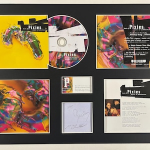 May include: A framed display of Pixies memorabilia, including an album, CD, and signed items. The album cover has a yellow background with a colourful abstract design. The CD is visible, along with a card and a signed note. The display is set against a black background.