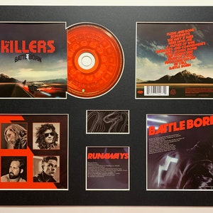 Puede incluir: Un set de CD enmarcado del álbum "Battle Born" de The Killers. La portada delantera presenta un diseño rojo y negro con el nombre de la banda y el título del álbum en letras grandes y en negrita. La portada trasera presenta una foto en blanco y negro de una persona que lleva una chaqueta de cuero con un diseño de rayo. El CD en sí es rojo con un centro negro.