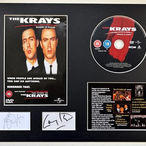 Peut inclure: Un cadre présentant la pochette du DVD The Krays, le disque lui-même et deux cartes d'autographes blanches. La pochette montre deux hommes en costume avec le texte "The Krays Bonded by Blood". L'ensemble inclut un bloc de texte et deux signatures.