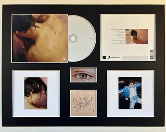 Op de afbeelding: Een ingelijste cd en foto's. De cd is wit en de foto's bevatten een close-up van een oog, een handtekening en afbeeldingen van de persoon in verschillende poses. De lijst is zwart met witte passe-partouts.
