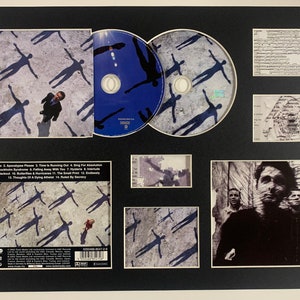 Puede incluir: Una presentación enmarcada del álbum "The Division Bell" de Pink Floyd con la portada del álbum, las letras y un CD. La portada del álbum presenta un fondo gris con siluetas de personas.