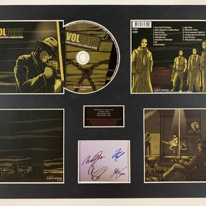 May include: A framed display featuring a Volbeat album, "Guitar Gangsters & Cadillac Blood." The display includes the album cover art, a CD, and a back cover with song titles. The artwork has a sepia tone with a man in a hat.