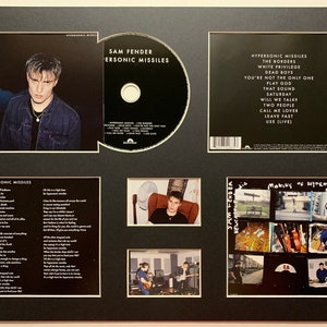 May include: A framed CD with a black and white photo of a man on the front cover. The CD is titled "Hypersonic Missiles" by Sam Fender. The back of the CD case lists the song titles. The frame also includes a photo of the artist and a collage of photos from the making of the album.