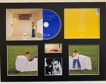 Op de afbeelding: Een ingelijste set foto's met een muzikant. De set bevat een foto van de muzikant in een kamer, een foto van de muzikant die op een bed zit, een foto van de muzikant die op een bed zit en een foto van de muzikant die op een bed zit. De set bevat ook een cd met de naam van de muzikant en de albumtitel. De albumtitel is "Harry's House". De cd is geel en heeft een lijst met nummers op de achterkant.