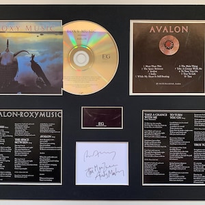 May include: A framed Roxy Music "Avalon" album display. It includes the album cover, CD, lyric sheets, and a signed autograph. The album cover features a dark, atmospheric image. The display is set against a black background.