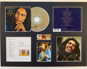 May include: A framed photo of Bob Marley with a CD and lyrics from his album "Legend". The photo shows Bob Marley in a black and white photo with a thoughtful expression. The CD is a gold disc with a black label. The lyrics are printed on a white sheet of paper. The frame is black with a gold border.