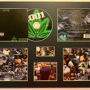 Può includere: Una presentazione incorniciata di un CD e una copertina di album per l'album 2001 di Dr. Dre. Il CD è di colore verde e nero con il numero 2001 in bianco. La copertina dell'album è nera con il titolo "Dr. Dre" e "2001" in bianco. La presentazione include anche un collage di foto dell'album.