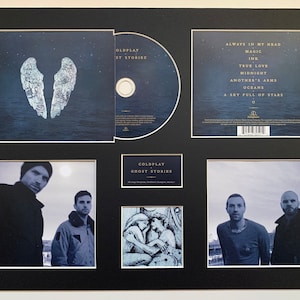May include: A black framed display with a CD, a CD case, and three photos. The CD case features a blue and white image of angel wings. The CD case back features the text "Coldplay Ghost Stories" and a list of songs. The CD is a silver disc with a blue and white image of angel wings. The photos are black and white and feature the band Coldplay. The photos are titled "Coldplay Ghost Stories".