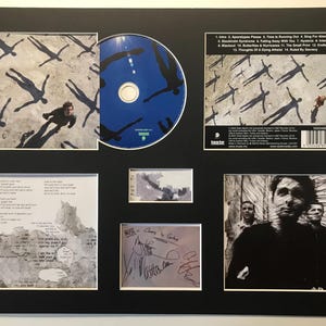 Op de afbeelding: Ingelijste cd en artwork met een band. De cd heeft een blauwe achtergrond met witte tekst. Het artwork bevat afbeeldingen van mensen en schaduwen, samen met handgeschreven handtekeningen. De lijst is zwart.