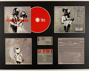Può includere: Un set incorniciato di copertine di album per l'album dei Blur "Think Tank". La copertina anteriore presenta un'immagine in bianco e nero di due persone con caschi da sub che si abbracciano. La copertina posteriore presenta un'immagine in bianco e nero di una persona con un casco da sub che tiene un bambino. Il CD è rosso con il nome della band e il titolo dell'album stampati in bianco.