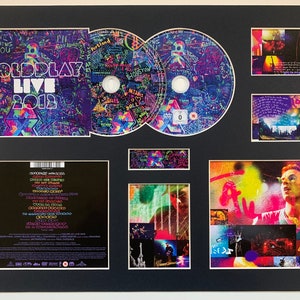 May include: A framed set of Coldplay Live 2012 DVD memorabilia. The set includes a DVD case with a colorful, graffiti-style design, two DVDs, and several photos of the band performing live. The DVD case features the text "Coldplay Live 2012" in a bold, white font.