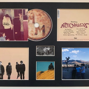 May include: A framed set of photos and a CD featuring the band Arctic Monkeys. The photos include the band members in various settings, including a desert landscape and a city street. The CD is titled "Humbug" and features the band's name in a stylized font.