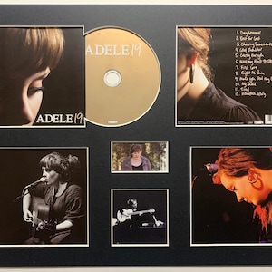 May include: A framed photo collage of Adele, a British singer-songwriter. The collage includes a CD, a photo of Adele on the cover of the CD, and several photos of Adele performing. The CD is titled "Adele 19".