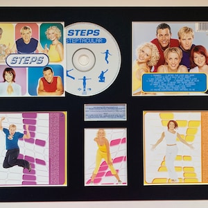 May include: A framed CD collection of the pop group Steps. The collection includes the album "Steptacular" with a CD, a back cover, and a front cover. The front cover features a group photo of the five members. The back cover features a list of songs on the album. The CD is in a clear plastic case. The frame is black with a white mat.