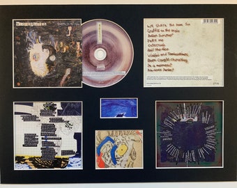 Può includere: Un set di CD incorniciato con sfondo nero e un passepartout bianco. Il CD è intitolato "Graffiti on the Train" di "Sece Ephonla". La copertina del CD presenta un design astratto in bianco e nero con un viso al centro. Il retro della custodia del CD presenta un elenco di titoli di canzoni. Il set include un CD, una custodia per CD e una stampa incorniciata della copertina del CD.