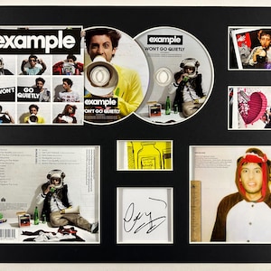 以下が含まれることがあります： アーティスト「Example」のCDとプロモーション資料を収めた額入りの音楽記念品。「Won't Go Quietly」というタイトルのCD。写真、アルバムアート、サインが黒い背景に配置されています。