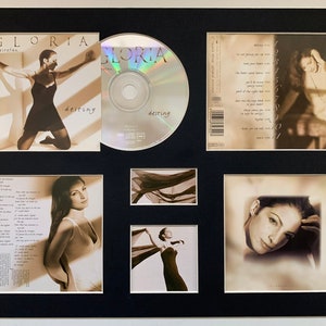 May include: A framed set of Gloria Estefan's "Destiny" album. The set includes the CD, the CD case, the back of the CD case, and three photos of Gloria Estefan. The photos are in black and white and sepia tones.
