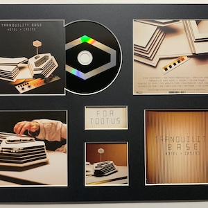 May include: A framed set of album artwork for the album "Tranquility Base Hotel & Casino" by Arctic Monkeys. The set includes a CD, a back cover, a front cover, and a close-up of a model of the hotel. The album title is printed on the front cover in white text on a black background. The back cover features a photo of the hotel with the album title and tracklist printed in white text on a black background. The close-up of the model shows a detailed view of the hotel's architecture.