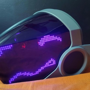 May include: A futuristic, gray and black helmet with a visor displaying a pixelated purple face. The helmet has a rounded top and a smooth, curved design, with a circular ear-like feature on the side. The visor glows with a purple light.