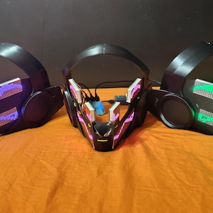 May include: Three black, futuristic-style masks with integrated LED lights. The masks have a sleek design with rounded ear pieces and a visor. The LED lights display various colors, including purple, green, and blue, creating a vibrant effect.