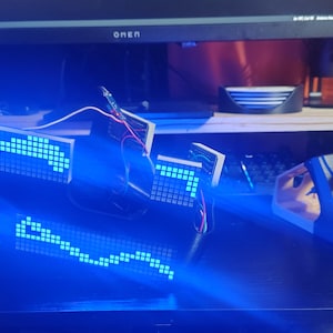 May include: A custom electronic device featuring multiple small LED dot matrix display modules connected by thin wires. The modules emit bright blue light in pixelated patterns, including a curved line and an arrow, set against a dark surface.
