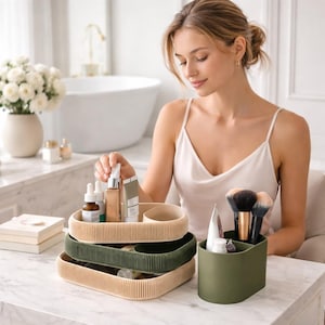 May include: A collection of beige and olive green cosmetic trays and a matching brush holder. The trays are stacked and hold beauty products. The items are on a white marble surface. The brush holder and trays are for bathroom organization.