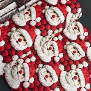 May include: A crochet blanket featuring a repeating pattern of Santa Claus faces with red hats and white beards, set against a background of small, textured red and white spheres. The Santa faces are crafted with detailed stitching and round noses.