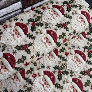 May include: A roll of cream-colored fabric featuring a repeating pattern of embroidered Santa Claus faces with red hats and white beards, interspersed with green holly leaves, red berries, and small gold stars throughout the design.