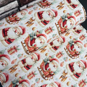 May include: A roll of fabric featuring a repeating pattern of Santa Claus figures, reindeer, gift boxes, and Christmas ornaments on a light background. The design includes Santa in a red suit, some riding in a sleigh, alongside small reindeer.