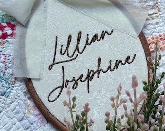 May include: An oval wooden embroidery hoop containing cream-colored fabric with a subtle white floral pattern. The name Lillian Josephine is embroidered in dark brown cursive thread. A sheer off-white ribbon is tied in a bow at the top.