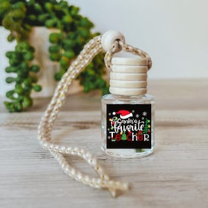 May include: A small, clear glass bottle with a wooden cap and a decorative rope. The bottle has a black label with the text "Santa's Favorite Teacher" in red, green, and white, with a Santa hat graphic. The bottle is a car air freshener.