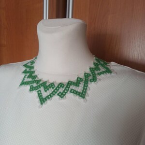 May include: A green geometric necklace with a white background. The necklace is made of small, square beads and has a zig-zag pattern. The necklace is displayed on a white mannequin head.
