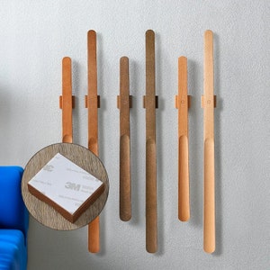 Peut inclure: Six crochets muraux en bois de différentes nuances de brun, fixés à un mur blanc. Les crochets sont conçus avec un sommet arrondi et une légère courbe en bas. Chaque crochet est fixé au mur avec une petite bande adhésive blanche.