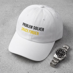 May include: A white baseball cap featuring the embroidered text "PROBLEM SOLVER" in black and "SNACK FINDER" in yellow on the front panel. The cap has a curved brim and a button on top. A silver-toned metal wristwatch with a black face is nearby.