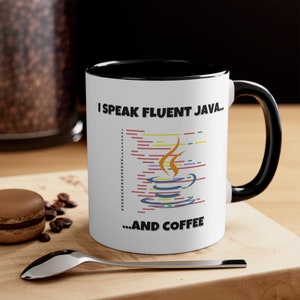 May include: White ceramic mug with a black rim and handle. The mug has a graphic of a coffee cup with steam and code, and the text "I speak fluent Java... ...and coffee".