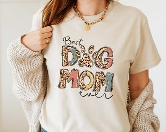 May include: A beige t-shirt with the text "Best Dog Mom ever" in colorful letters with a paw print in the middle of the word "Dog". The letters are decorated with a leopard print pattern.