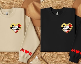 Può includere: Due felpe con una grafica di Topolino e Topolina che si baciano sul davanti. Le felpe sono beige e nere. Le maniche hanno un cuore rosso con le lettere "M" e "R" su di esse.