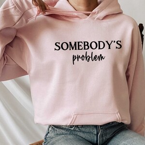 Op de afbeelding: Een roze hoodie met de tekst "SOMEBODY'S problem" in zwarte letters.