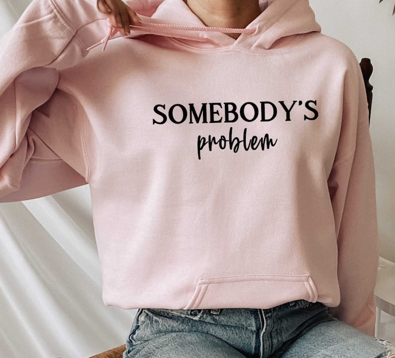 Op de afbeelding: Een roze hoodie met de tekst "SOMEBODY'S problem" in zwarte letters.