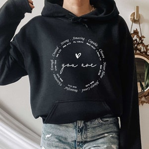 May include: Black hooded sweatshirt with a white circle graphic that says "You are" with a heart in the center. The circle is surrounded by words like "Strong", "Amazing", "Capable", "Chosen", "Never Alone", "Always Loved", "Beautiful", "Victorious", "Enough", "Created".