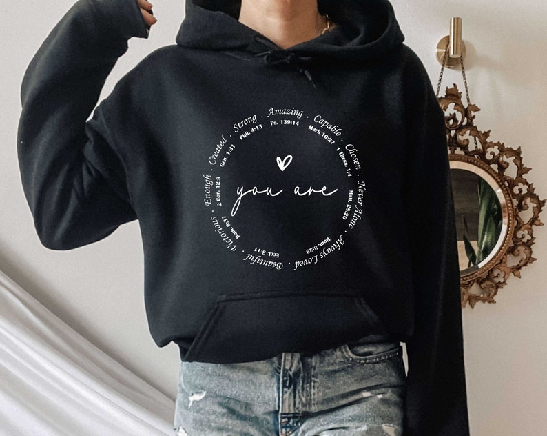 May include: Black hooded sweatshirt with a white circle graphic that says "You are" with a heart in the center. The circle is surrounded by words like "Strong", "Amazing", "Capable", "Chosen", "Never Alone", "Always Loved", "Beautiful", "Victorious", "Enough", "Created".