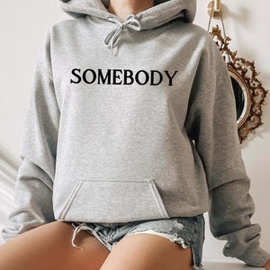 Op de afbeelding: Een grijs hoodie met het woord "SOMEBODY" in het zwart op de voorkant.
