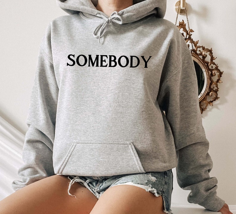 Op de afbeelding: Een grijs hoodie met het woord "SOMEBODY" in het zwart op de voorkant.
