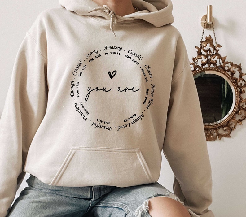 May include: A beige hoodie with a black circle graphic that says "You are" with a heart symbol and a list of positive affirmations around the circle. The affirmations include "Strong", "Amazing", "Capable", "Chosen", "Never Alone", "Always Loved", "Beautiful", "Victorious", "Enough", and "Created".